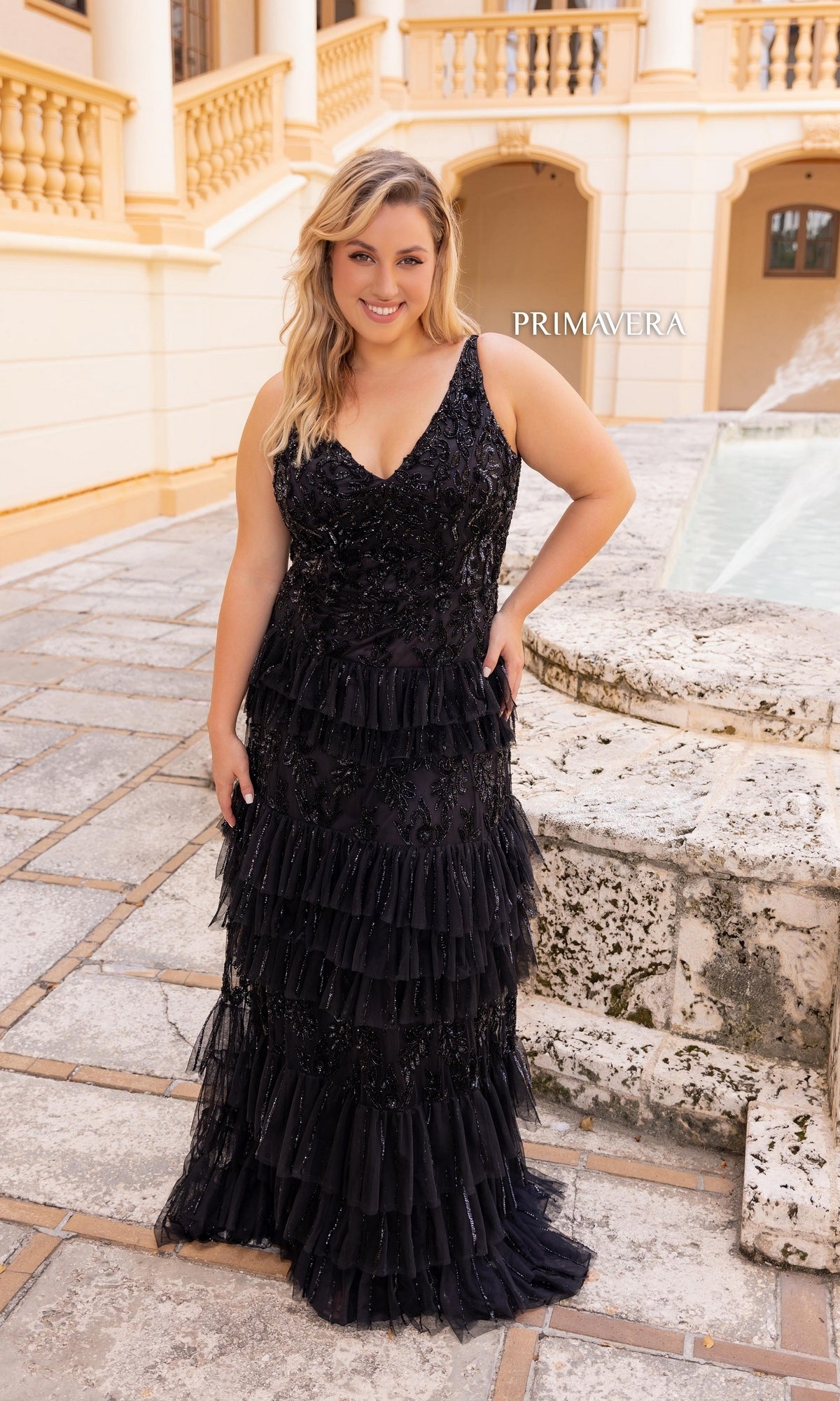 A woman, smiling at the camera, stands outdoors on a stone patio near a fountain wearing the Primavera Tiered Plus-Size Long Prom Dress 14052 in black.